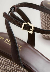 Sandalo marrone con texture intrecciata sulla suola, cinturini in plastica e una fibbia color oro. Presenta un tacco alto e una soletta marrone con marchio.