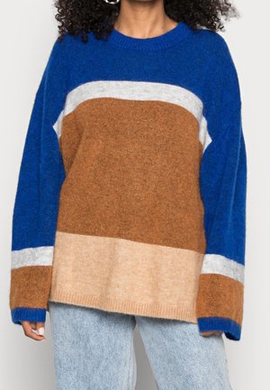 Femme portant un pull duveteux à blocs de couleurs bleu, marron, beige et blanc avec des manches longues et un jean bleu clair, debout devant un fond uni.