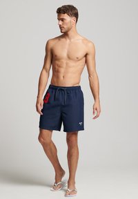 Costumi da bagno blu navy con coulisse, caratterizzati da un numero rosso "8" sul lato sinistro e un logo in basso a destra. Infradito completano l'outfit.