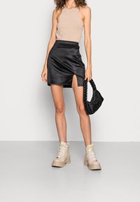 Top halter beige, falda mini de satén negra con apertura lateral, usando botas gruesas beige, sosteniendo un bolso trenzado negro.