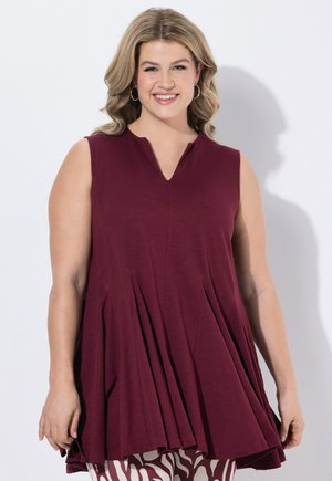 Frau mit welligem blondem Haar lächelt, trägt ärmellose burgunderfarbene Tunika mit Falten und gemusterte Hose, steht vor weißem Hintergrund.