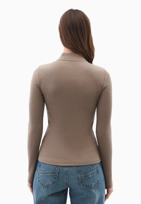 Kvinde med langt brunt hår iført en tætsiddende taupe ribbet langærmet top og blå denimjeans, set fra ryggen på en hvid baggrund.