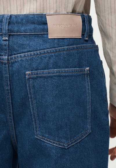 Λεπτομέρεια πίσω τσέπης μπλε τζιν με εμφανείς ραφές και μπεζ ετικέτα Maison Kitsuné Paris πάνω από τη μέση της ζώνης.