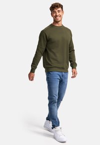Sudadera verde olivo con cuello redondo y puños acanalados, combinada con jeans ajustados azul claro y zapatillas blancas. Textura suave, diseño casual.