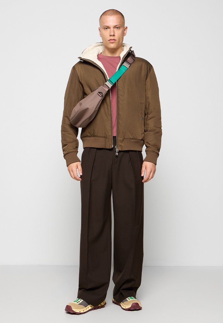 Veste bomber marron avec col en fourrure, portée sur une chemise bordeaux. Pantalons larges marron, sneakers marron et beige, et un sac bandoulière marron.