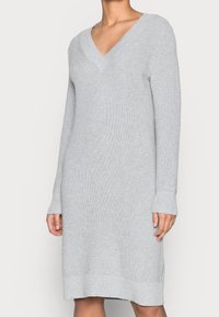 Femme portant une robe tricotée à manches longues de couleur gris clair avec un décolleté en V et des poignets et ourlet côtelés, debout devant un fond uni.