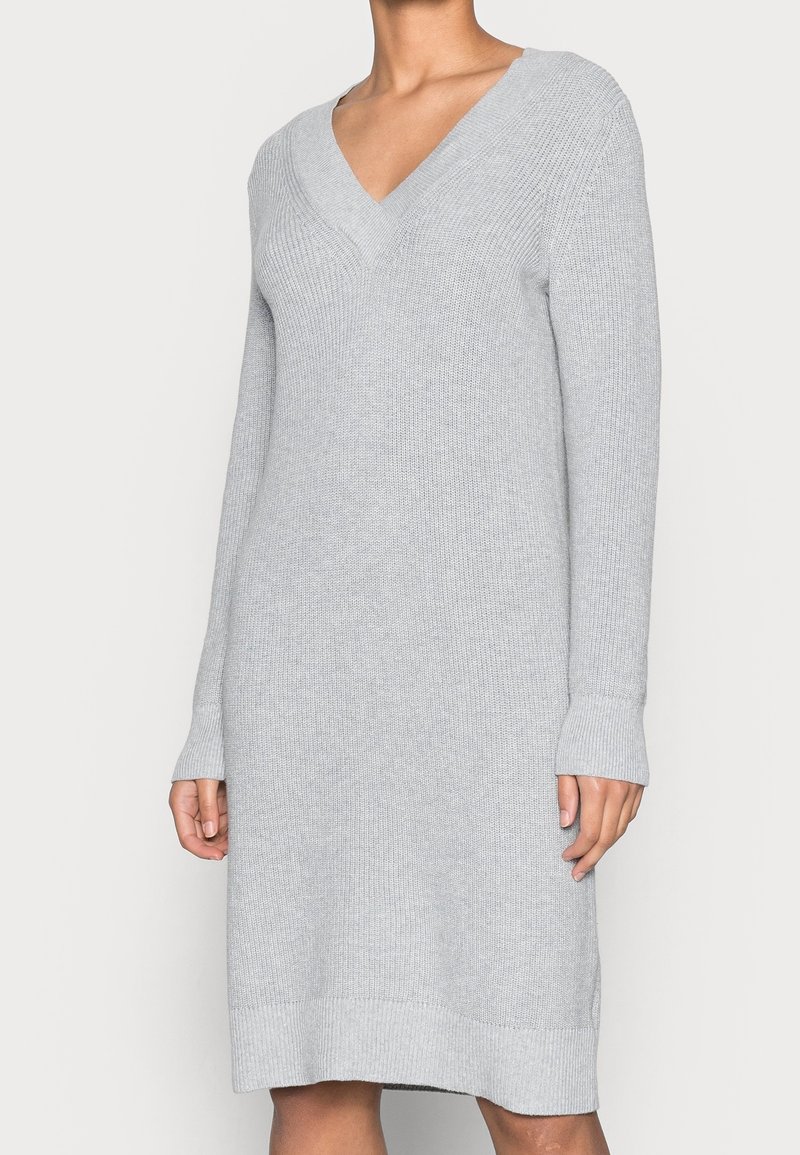 Femme portant une robe tricotée à manches longues de couleur gris clair avec un décolleté en V et des poignets et ourlet côtelés, debout devant un fond uni.