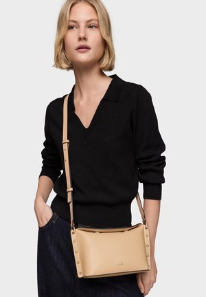 Kobieta ubrana w czarny sweter z dekoltem w serek i ciemne spodnie, niosąca beżową torbę na ramię z detalami w postaci ćwieków i wytłoczonym logo marki.