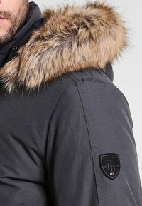 Homme portant une veste gris foncé avec capuche doublée de fourrure et un patch logo Hilfiger sur le haut du bras, visible de l'épaule au menton.