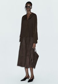 Donkerbruin, losvallend top met een kraag en plooien, gecombineerd met een bijpassende midi-rok, zwarte slingback schoenen en een grijze tas.