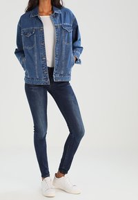 Chaqueta de denim en azul medio con un corte holgado, dos bolsillos en el pecho y cierre de botones. Combinada con jeans skinny oscuros y zapatillas blancas.
