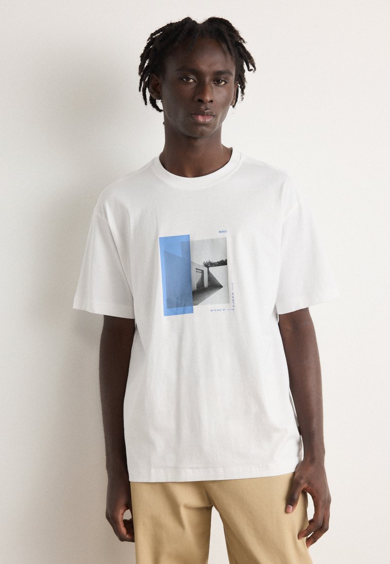 Balta medvilnė t-shirt marškiniai su mėlyna ir pilka grafika, vaizduojančia geometrines formas ir architektūrinius elementus. Trumpomis rankovėmis, laisvas kirpimas.