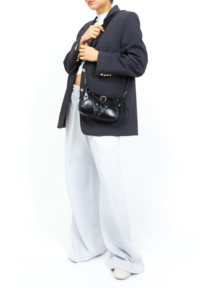 Borsa a tracolla in pelle nera con forma curva, caratterizzata da borchie decorative e un dettaglio con fibbia, abbinata a un blazer grigio oversize e pantaloni chiari.