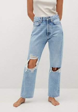 Femme portant un jean bleu clair taille haute déchiré aux genoux, debout pieds nus sur un fond blanc uni.