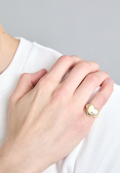 Zelta gredzens sirds formā, ar gludu virsmu un mazu dārgakmeņu akcentu. Valkāts uz rokas ar gaišu ādu uz balta fona.