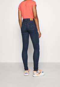 Jeans de mezclilla oscuros de corte ajustado y cintura alta, con bolsillos traseros sutiles y costuras claras, combinados con una blusa naranja de manga corta y zapatillas.