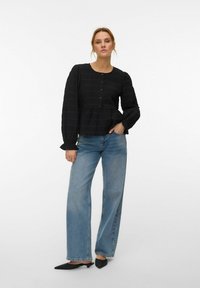 Femme debout, une main dans la poche, portant un chemisier noir boutonné, un jean bleu à jambes larges et des ballerines noires pointues sur fond blanc.