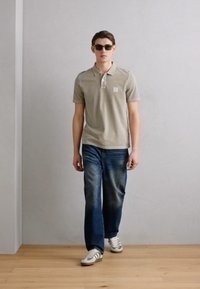 Lys beige polo skjorte med krage og logo, sammen med blå denim jeans og hvite sneakers. Modellen har på seg mørke solbriller og står innendørs.