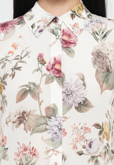 Bluse mit floralem Muster, die einen weißen Hintergrund mit verschiedenen Blumen in Pink, Lila und Gelb zeigt. Sie hat einen spitzen Kragen und eine Knopfleiste.