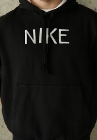Svart hoodies i bomull med framficka. Har fet vit "NIKE" text på bröstet och en hood med dragsko.