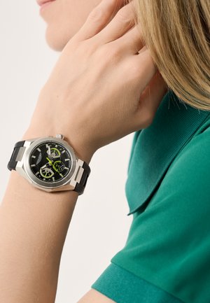 Silberne Edelstahluhr mit schwarzem Kautschukarmband. Sie verfügt über ein schwarzes Zifferblatt mit silbernen und grünen Akzenten, einschließlich Chronographen-Unterzifferblätter.