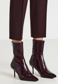 Stivali alla caviglia lucidi di un borgogna scuro con punta appuntita, cerniera laterale e tacchi alti sottili, indossati con pantaloni cropped abbinati.