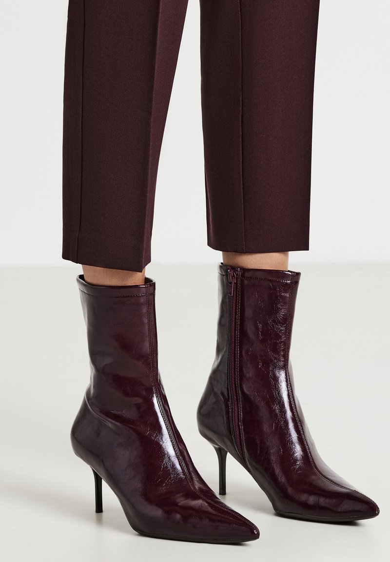 Stivali alla caviglia lucidi di un borgogna scuro con punta appuntita, cerniera laterale e tacchi alti sottili, indossati con pantaloni cropped abbinati.