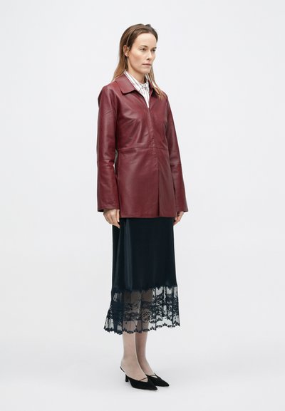 Γυναίκα που στέκεται φορώντας ένα μπορντό δερμάτ�ινο μπουφάν πάνω από μια ριγωτή μπλούζα και μαύρη σατινέ φούστα με δαντελένιο τελείωμα, φορώντας μαύρα μυτερά τακούνια.