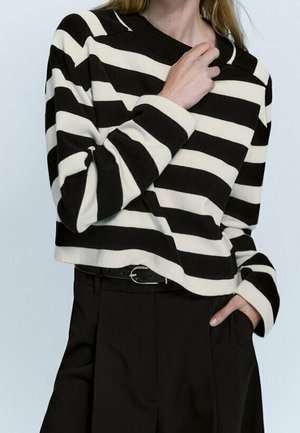 Femme portant un pull à manches longues à rayures horizontales noires et blanches, rentré dans un pantalon noir taille haute avec une ceinture.