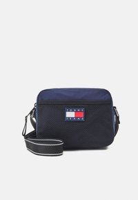 Marinblå tvärsöver axeln-väska med nätstruktur, med en logotypapplikation och en justerbar rem med vit sömnad. Rektangulär design.