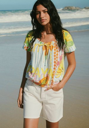 Young woman with long dark hair wearing a colorful short-sleeve blouse and white shorts standing on a sandy beach near the ocean.
