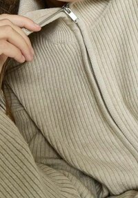 Beige ribbad tröja med hög krage och silverblixtlås. Tyget verkar mjukt med ett strukturerat mönster och avslappnad passform.
