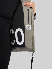 Grå quiltet jakke med sort for og et stort hvidt motiv. Indeholder en lynlåslomme og en label med teksten "En særlig kram lavet af stof."