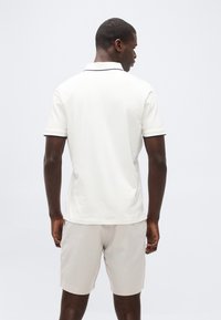 Homme portant un polo blanc à manches courtes et un short beige clair, debout de dos contre un fond uni clair.