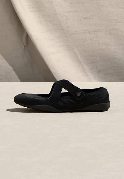 Μαύρο επίπεδο παπούτσι με σταυρωτό λουράκι και λεπτομέρεια με κουμπί, σχεδιασμένο για καθημερινή χρήση, τοποθετημένο σε μπεζ επιφάνεια με ουδέτερο φόντο από ύφασμα.