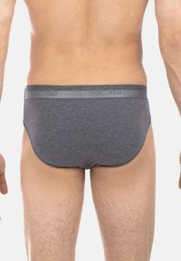 Mann in schlichten grauen Unterhosen, der mit dem Rücken zur Kamera vor einem weißen Hintergrund steht.