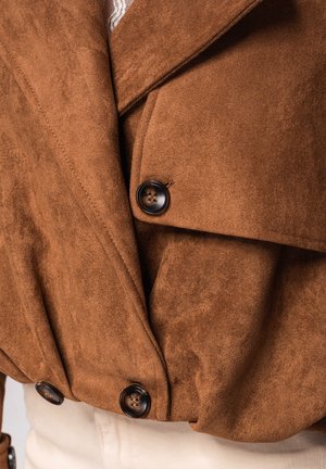 Veste en suède marron avec de grands boutons foncés et de larges revers, portée sur un pantalon de couleur claire.