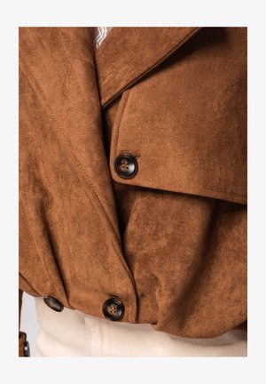 Veste en suède marron avec de grands boutons foncés et de larges revers, portée sur un pantalon de couleur claire.