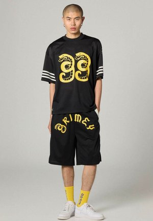 Jonge man gekleed in zwarte sportkleding met een geel slangontwerp en het cijfer "99" op het shirt, de tekst "Grimey" op de short, gele sokken en witte sneakers.
