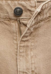 Gros plan d’un jean en denim beige avec une fermeture éclair partiellement ouverte et un bouton métal noir portant l’inscription "Swing Jeans Established 1984."