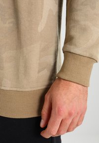 Beige genser med en subtil kamuflasjemønster, ribbestrikkede mansjetter og en myk tekstur, med detaljer ved sidesømmen og en avslappet passform.