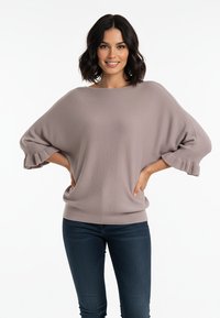 Oversizowy sweter w jasnym odcieniu taupe z fakturowanym splotem, szerokim dekoltem w łódkę i bufkami na rękawach, zestawiony z dopasowanymi ciemnoniebieskimi jeansami.