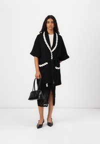 Robe noire de style kimono avec des accents blancs en forme de coquille, nouée à la taille, associée à une jupe noire et des talons aiguilles noirs, tenant un sac à main.