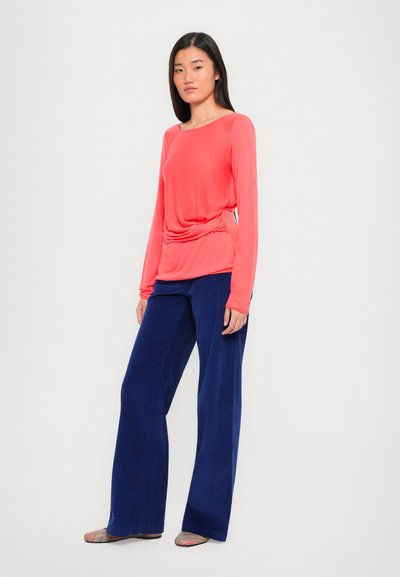Top a maniche lunghe in corallo con dettaglio arricciato sui lati, abbinato a pantaloni larghi blu navy realizzati in tessuto morbido. Il modello indossa scarpe piatte a righe.
