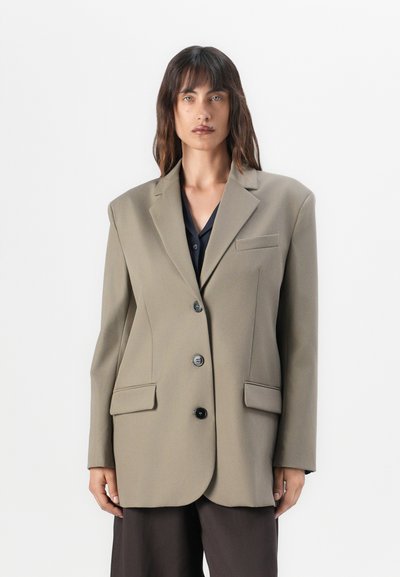 Oversized beige blazer with notched lapels, two front pockets, and three black buttons. Made of smooth fabric, features a structured shape.