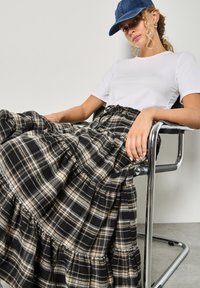 Gonna a quadri nera e beige con design a strati, realizzata in un tessuto testurizzato, abbinata a una maglietta bianca semplice e un cappellino di denim.