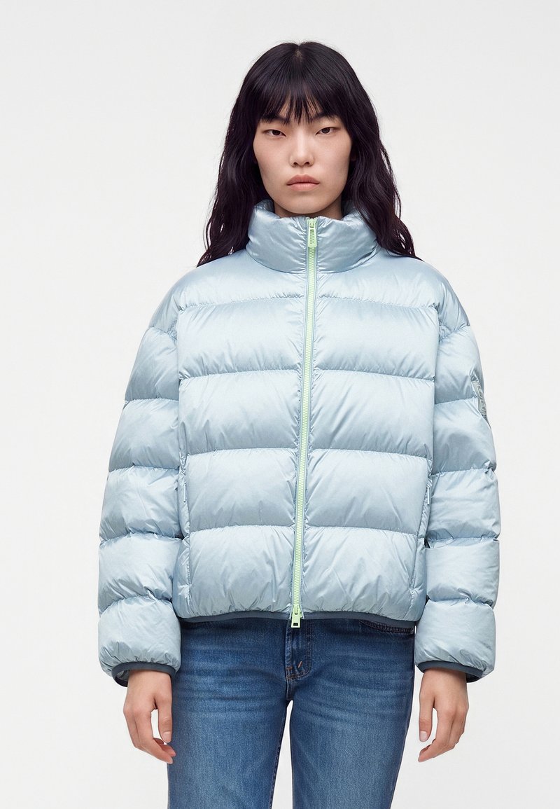 Femme aux longs cheveux foncés porte une doudoune bleu clair zippée, associée à un jean bleu, debout devant un fond uni.