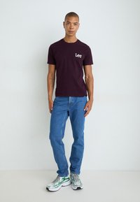 Burgunderfarvet bomulds t-shirt med "Lee" logo, parret med lyseblå denim jeans og sneakers med grønne accenter. Model står foran en neutral baggrund.