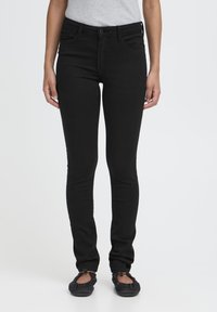 Frau trägt schwarze Skinny-Jeans und schwarze Ballerinas, steht mit entspannten Armen neben dem Körper vor einem schlichten Hintergrund.