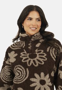Donna con capelli scuri che indossa un pullover di pile marrone con motivi floreali e spirali beige, sorridente e che guarda a destra.